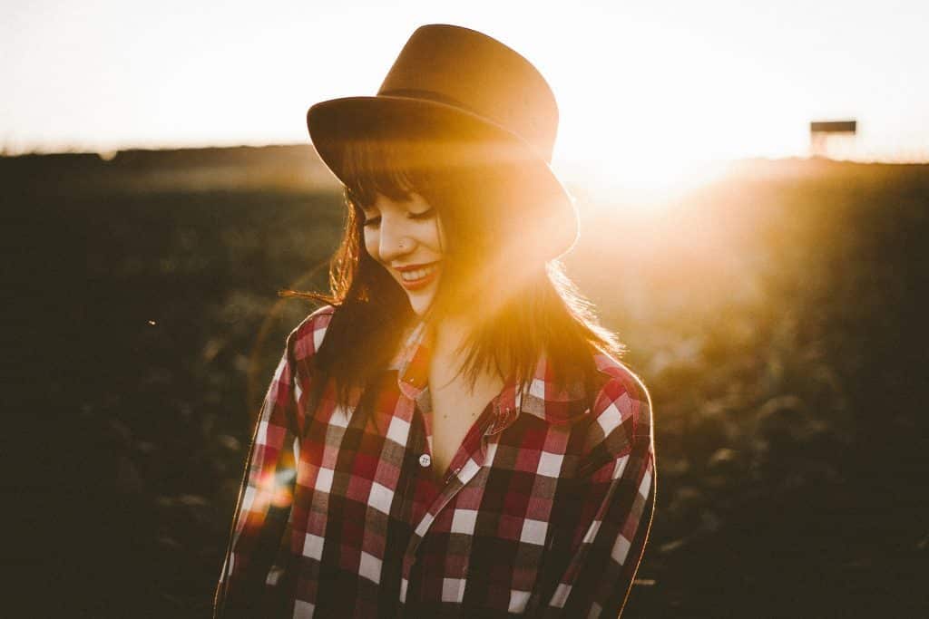 4 ways mindfulness enhances happiness: Woman wearing a hat and checkered shirt stands in the sun smiling