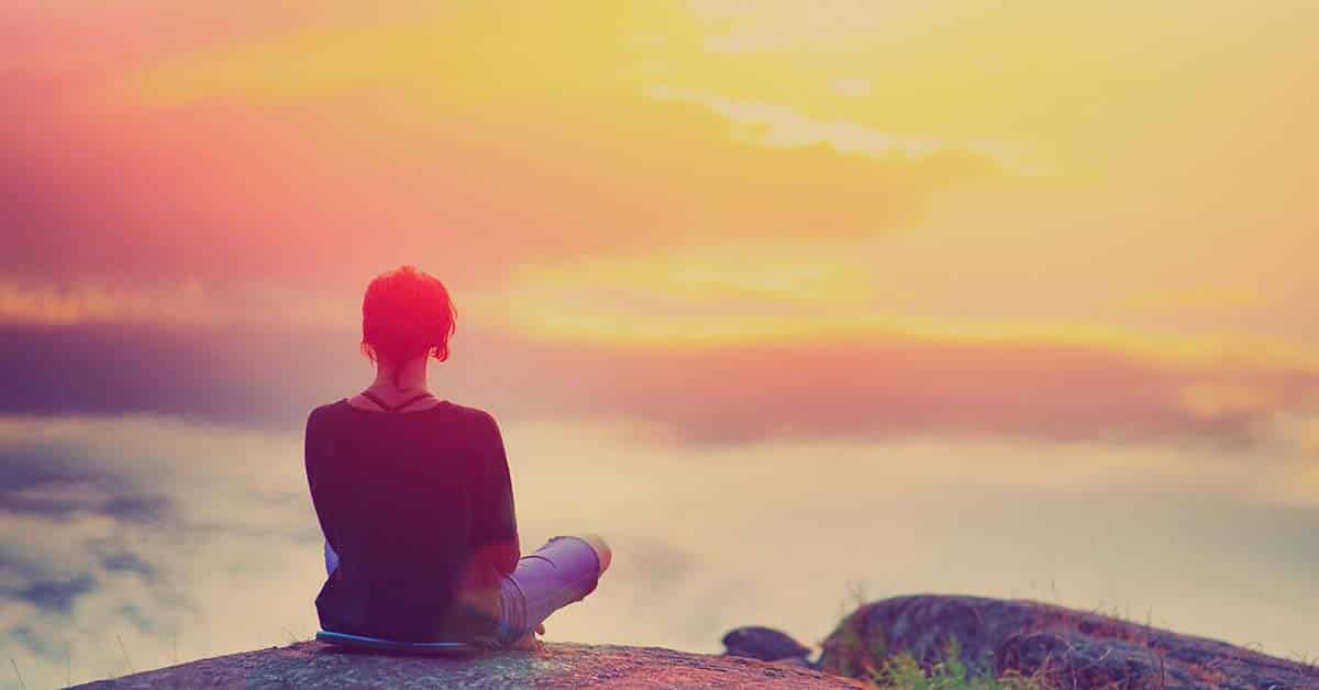 Young woman sitting enjoying peaceful moment of beautiful colorful sunset. In the reflection of the lake water sees clouds and sun. Vintage mood, concepts of winner, freedom, happiness.